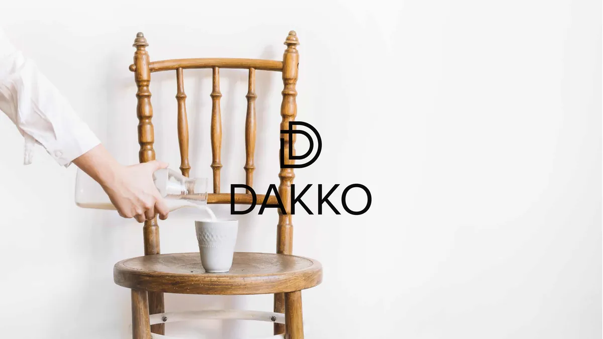Dakko: presentation cover, a hand pouring milk into a cup on a wooden chair, black DAKKO logo