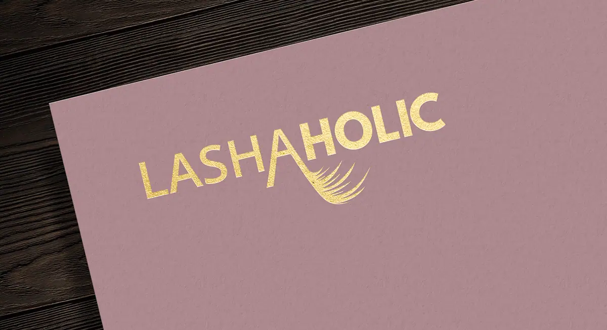 Lashaholic: złote logo na pudrowo-różowym papierze na ciemnym drewnie